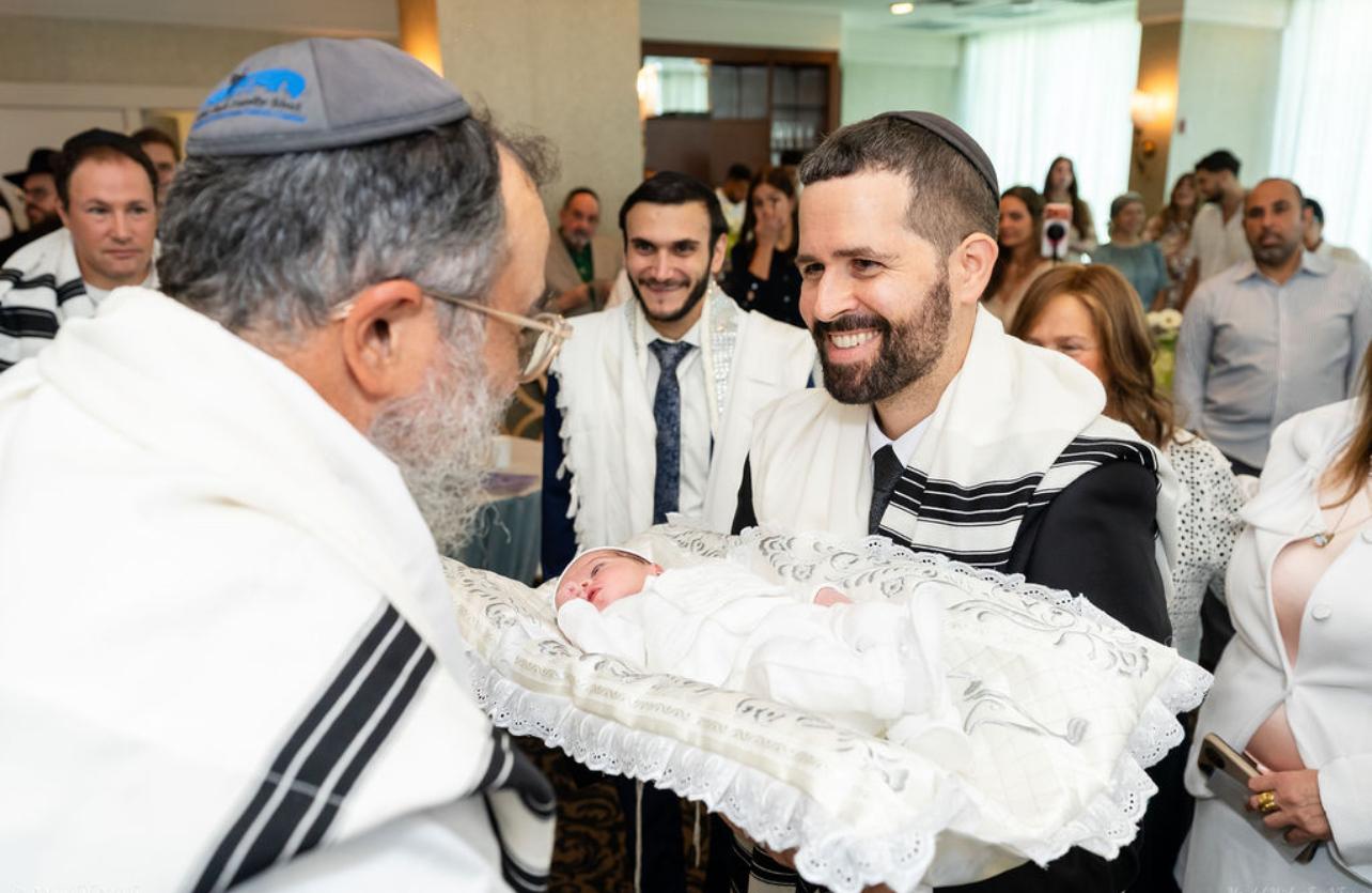 Rabbi Yossi Srugo smiling as he hands a newborn baby to the sandek during a Brit Milah ceremony in Weston